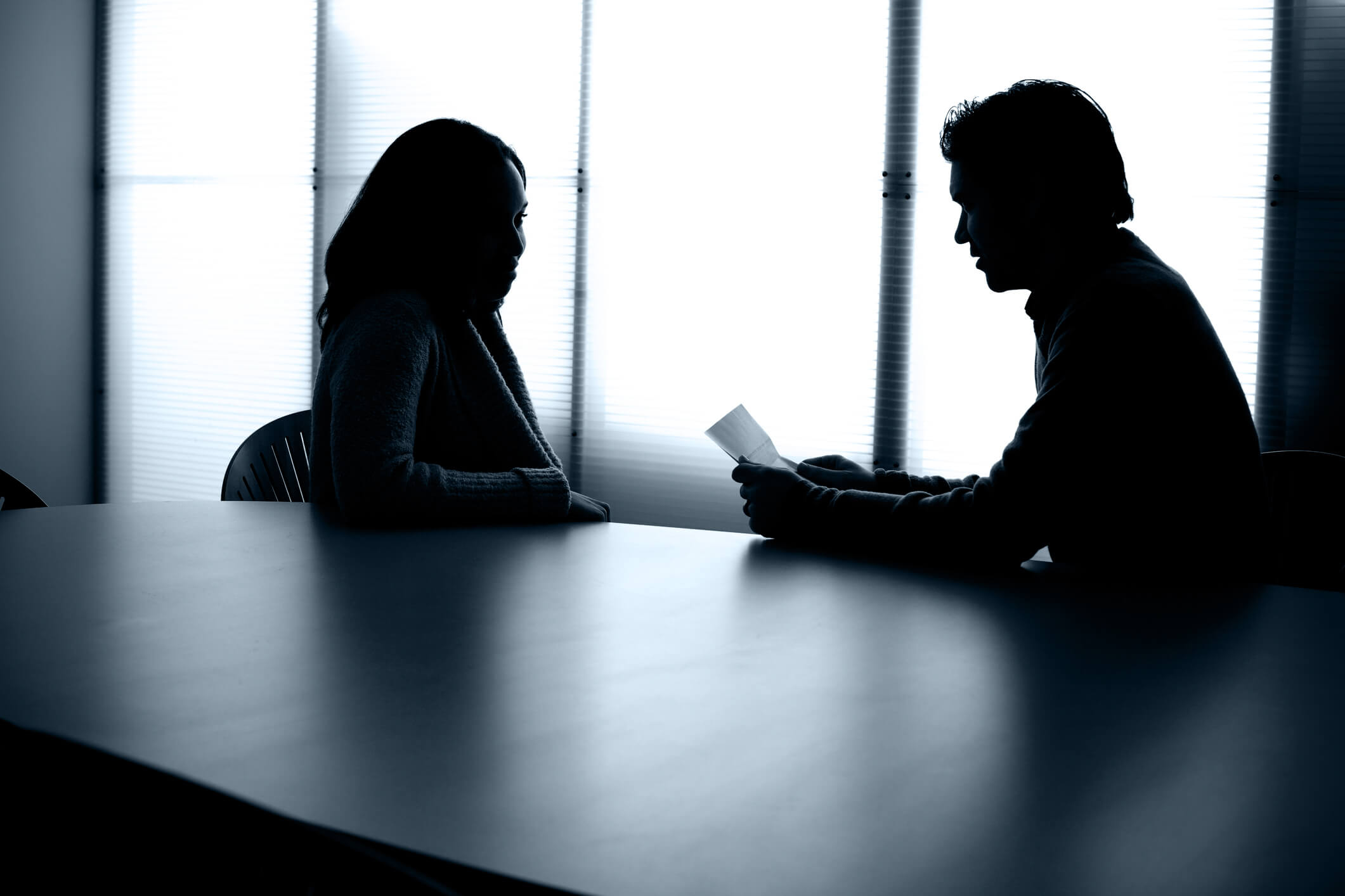 Two back lit people,one female, one male, in near silhouette, sit at a conference table having a discussion in a modern office environment.