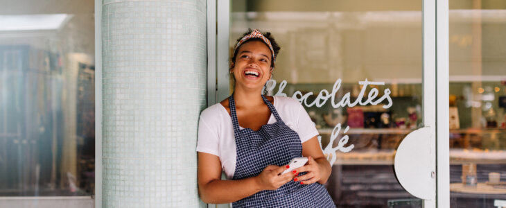 Successful cafe owner, a young woman, stands in her restaurant using a smartphone. Happy female entrepreneur using technology to manage her small business efficiently.